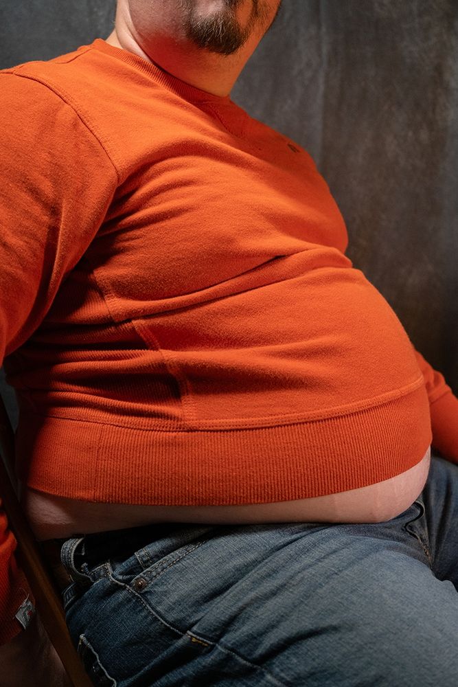 Babyblueboxers sitting in a chair with an sweater shirt and jeans, belly is hanging out below the shirt.