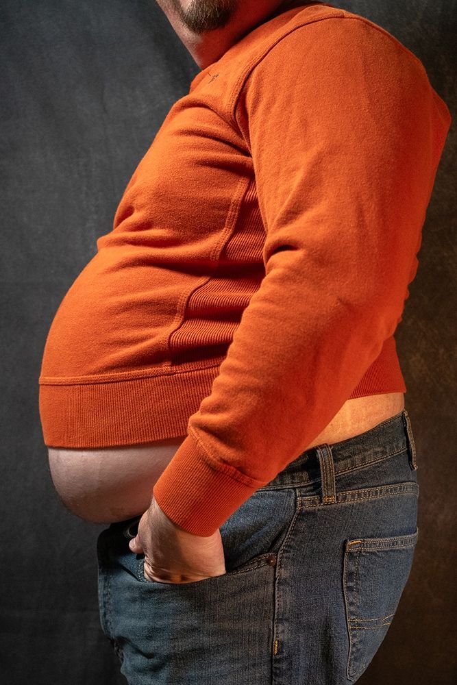 Babyblueboxers is standing to the side in an orange sweater and jeans, belly is hanging out below the shirt.