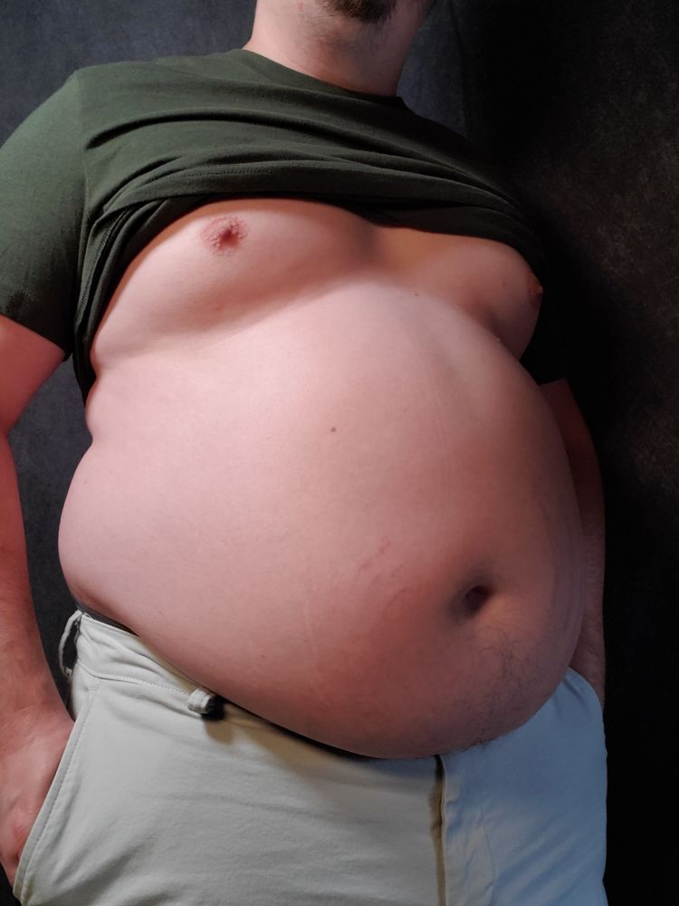 Babyblueboxers standing in a green shirt and khaki shorts. Belly is round and out, turned slightly to the side. 