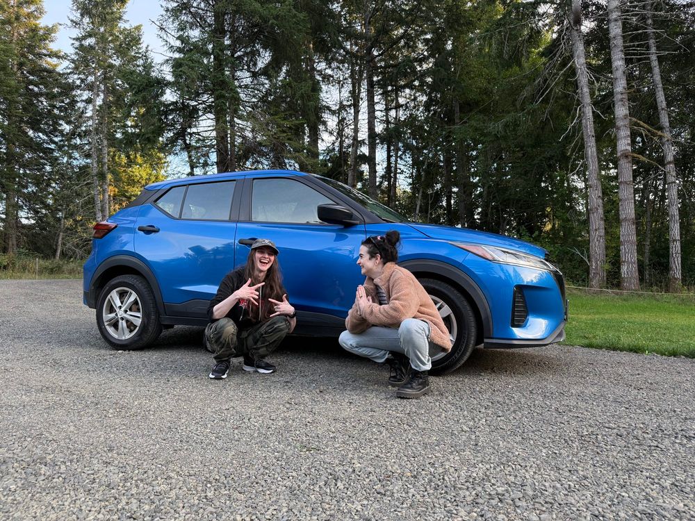 A couple crouched in front of the side of a blue Nissan Kicks. The person on the left in camo pants, a black hoodie, and a hat is holding up hand signs that are something like if the sign for love and the rock and roll sign had a baby. The person on the right in a fuzzy tan coat and light blue jeans with black hair has his hands in the preaching sign. They are looking at each other and laughing and deeply in love.
