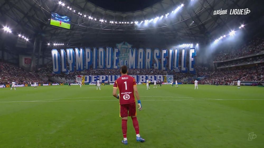 Au premier plan, Rulli de dos et en arrière plen le Virage Sud avec une immense voile montée « Olympique de Marseille » et un message en bas du Virage « depuis 1899 »