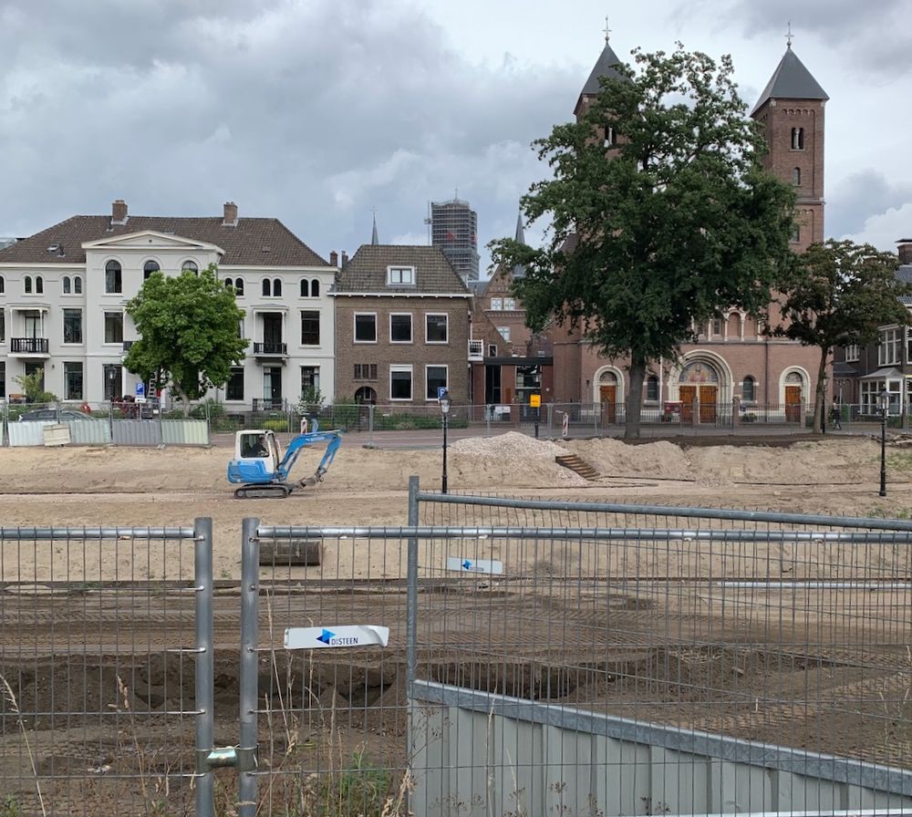Sand field with small excavator, construction fencing. In the background facades of buildings, a tree.
