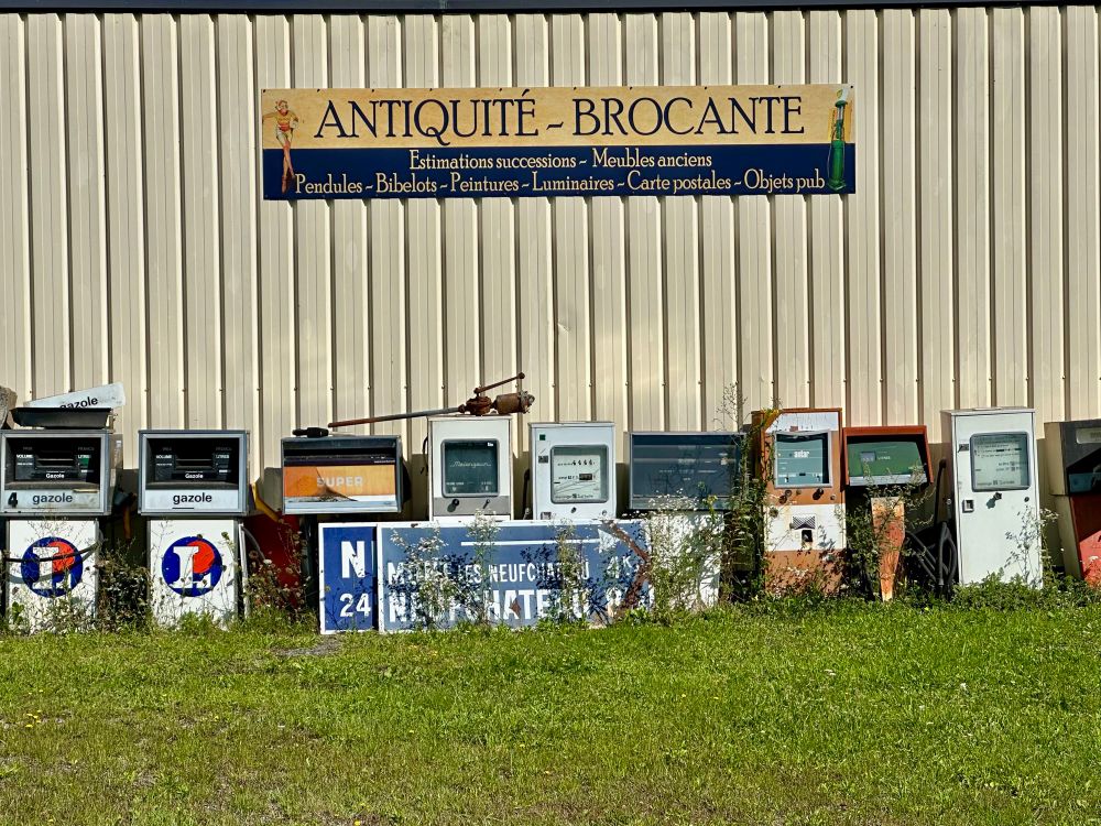 Old fuel pumps outside against a wall. On top of them is a sign with the text 'Antiquité - Brocante' (Antiques - Flea Market).