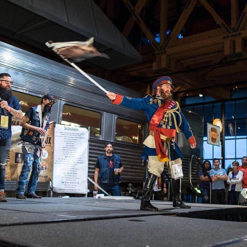 Regev in Sacramento from 2013, with a freestyle beard and a bear flag-esque flag. He is posed in Soviet military surplus gear because of course he is. 