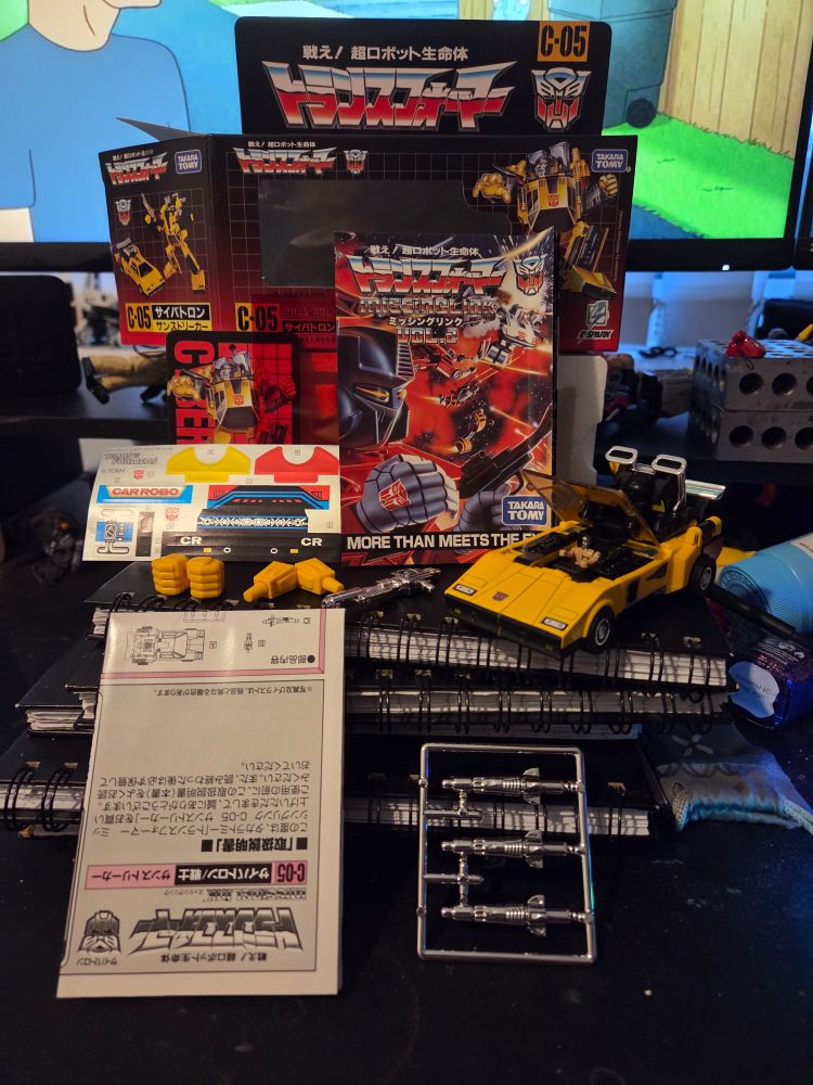 Transformers Missing Link Sunstreaker. The box, instructions, stickers, and extra parts sit next to a yellow lamborghini with a micro man in it.
