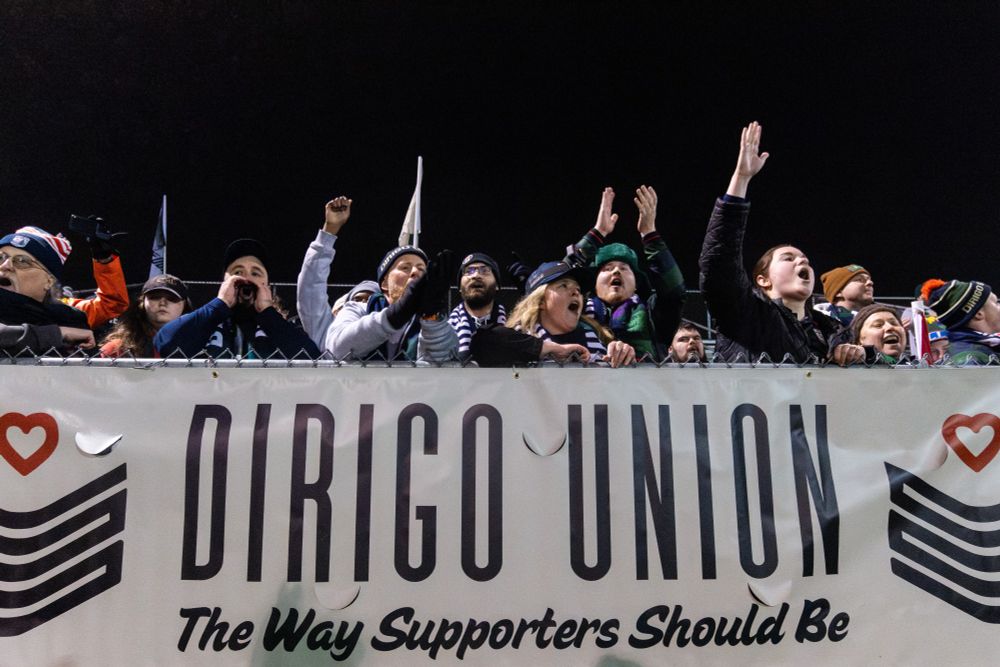 Dirigo Union supporters cheering on Hearts of Pine in Lewiston, Maine on 3/20/25 in front of a banner that reads "Dirigo Union. The way supporters should be."