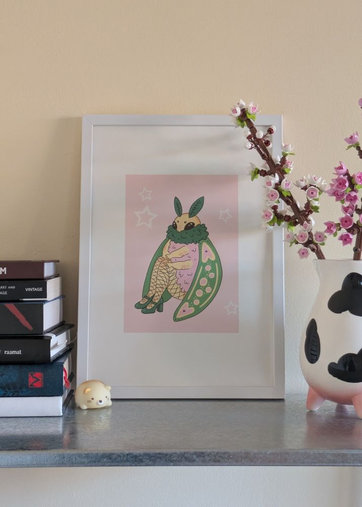 Print of an moth illustration. The moth has long legs in fishnets and green chonky high-heels. It has a pink fur. The moth is sitting on the ground and has its  hands on its knees. The print is on a metallic shelf in a white picture frame. On the left of the frame is a stack of books and a little cat figurine. On the right is a carafe that has a cow print and its legs are pink udders. In the carafe are two lego cherry blossom flowers.
