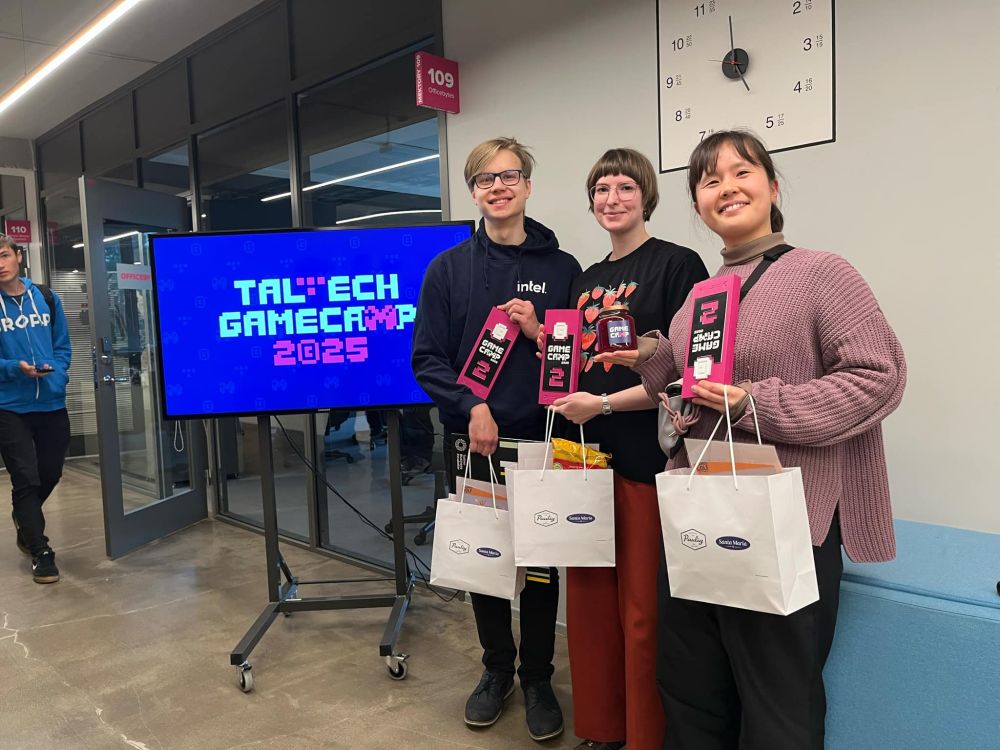 Picture of 2 women and 1 guy standing and holding gift bags and a 2nd place trophy. Next to them is a TV displaying text "Taltech gamecamp 2025"