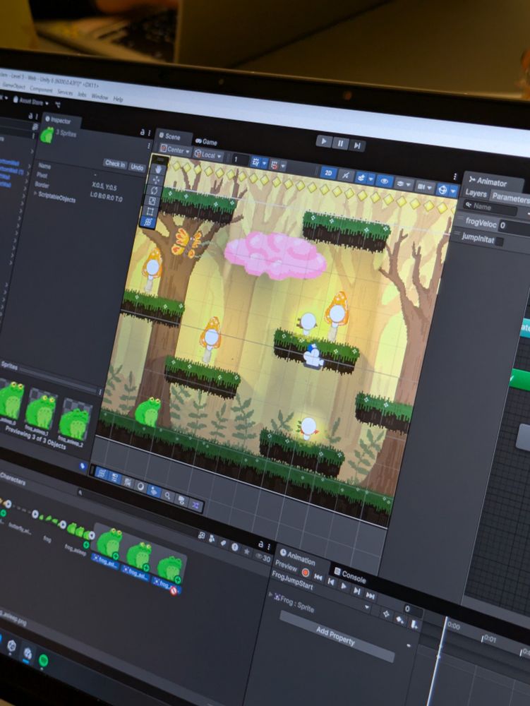 Picture of a laptop screen: Unity program is open. You can see a platformer game with warm cozy lightning. There's a frog, a butterfly, ponk cloud and 3 glowing mushrooms.