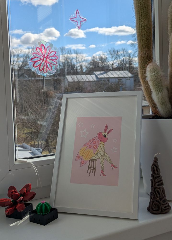 original post photo frames in a white picture frame. Next to it is a candle and two lego plants. on the background is a big cactus and a window where you can see a blue sky.