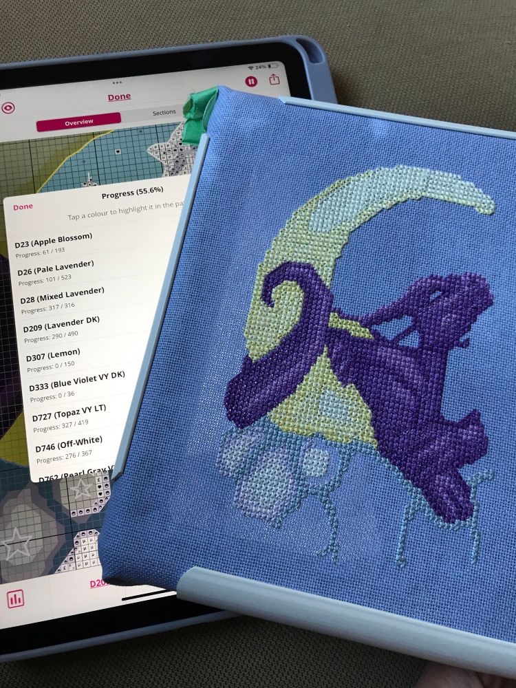 A partially finished cross stitch project sits atop an iPad displaying progress at almost 56%.