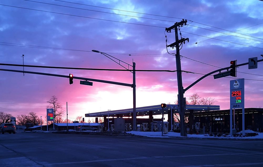 A pale yellow, pink, purple, and blue cloud sky at sunrise with a gas station sillouette
