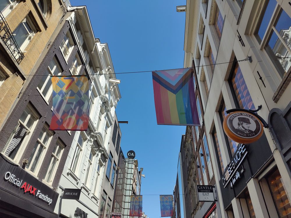 Eine Pride-Flagge ist neben einer bunten Flagge mit der Aufschrift "750" an einem Seil über eine Straße gespannt. Im Hintergrund hängen weitere solche Flaggen.