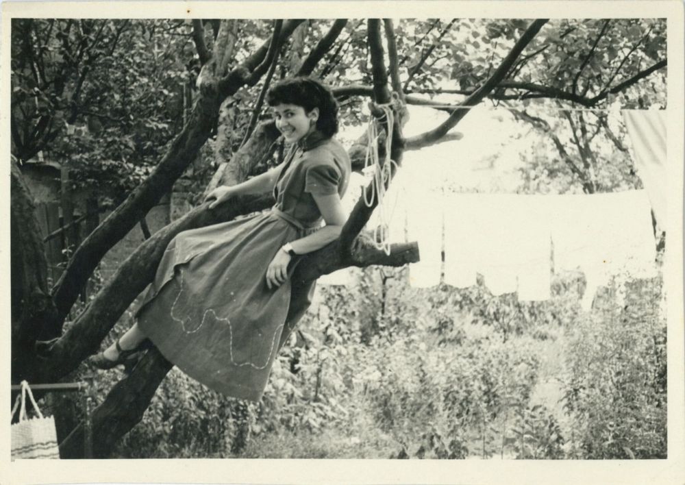 A photograph of a young woman in a tree smiling to the camera. She is wearing a long dress and shoes with large heels not well suited to climbing trees. To her right are clothes on a washing line. Perhaps she has climbed a tree in a garden to mark a special occasion. The image is from France circa 1950.