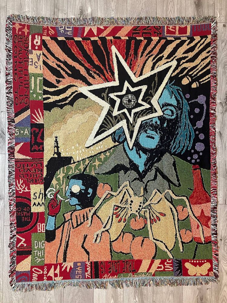 Picture of a woven blanket / tapestry with a design inspired by Disco Elysium. Harry Du Bois cries a single tear while Kim Kitsuragi lifts a hand in the fashion of Dolores Dei. The borders are kind of collage style. There is a lot of red.