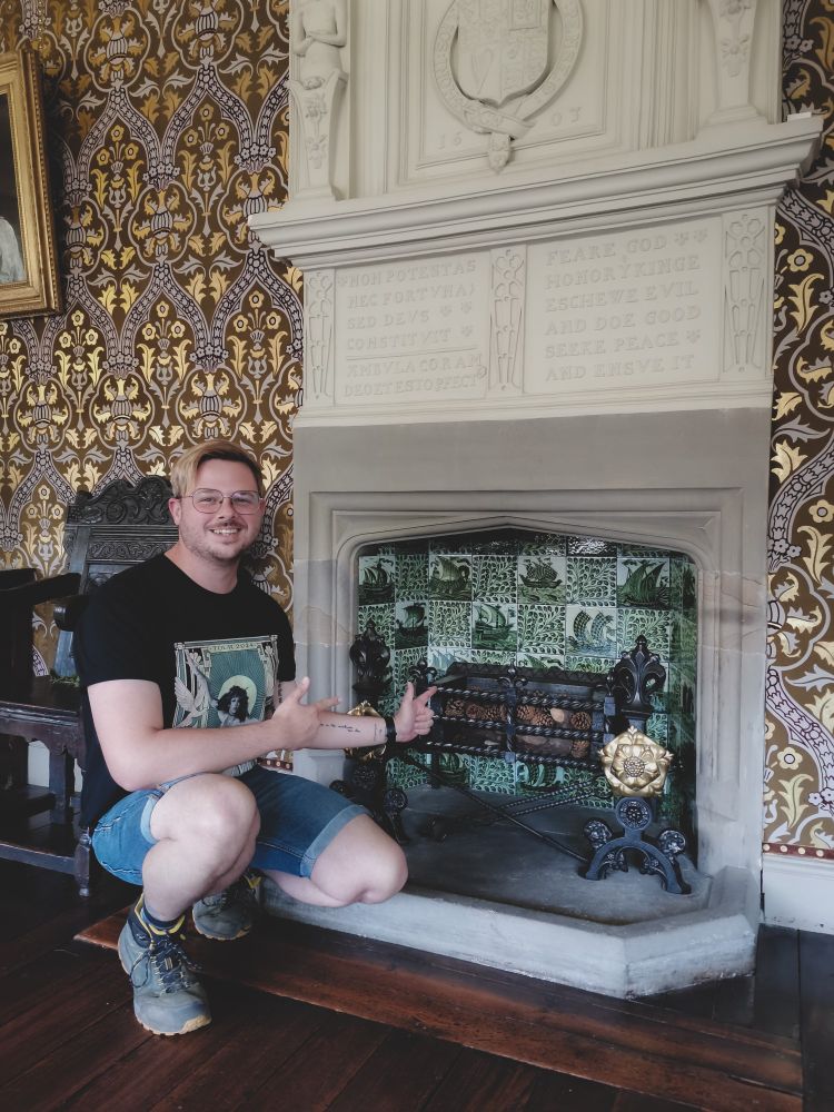 Steven crouched in front of a stome fireplace which is tiled with the William De Morgan Green Galleon design. 