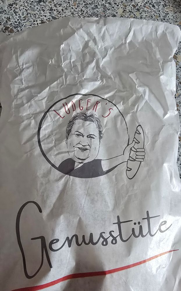 Bäckerei Brötchentüte. Darauf dass gezeichnete Bild eines Mannes, der ein Baguette hält. Darauf steht: Ludger's Genusstüte.