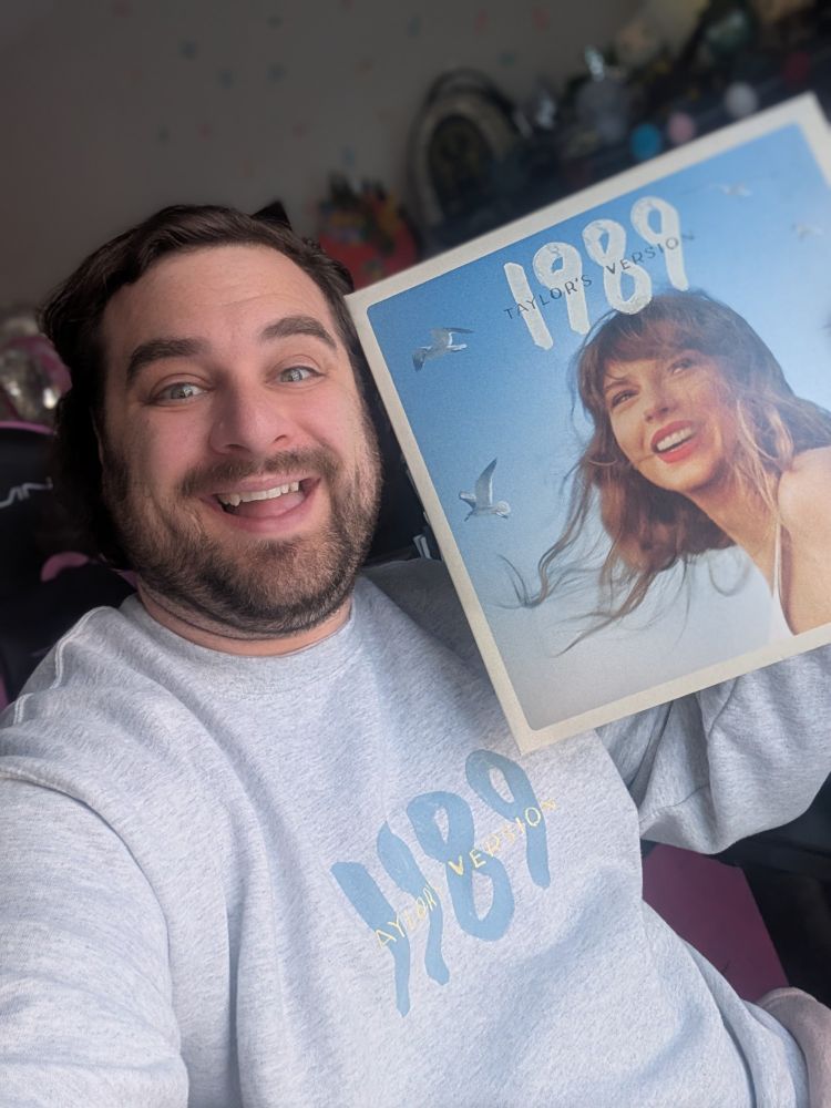 White male with brown hair and big smile wears Taylor Swift 1989 sweatshirt while holding the 1989 vinyl