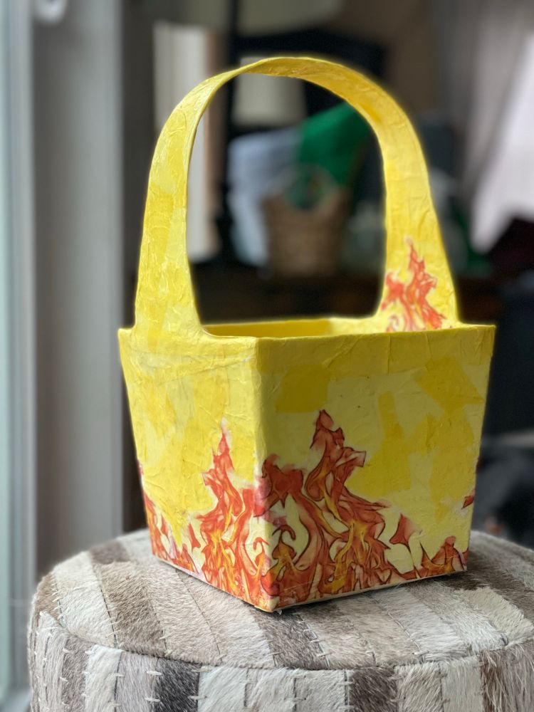 Square paper-covered basket with handle. Yellow, decorated with orange flames. The proverbial hand basket.