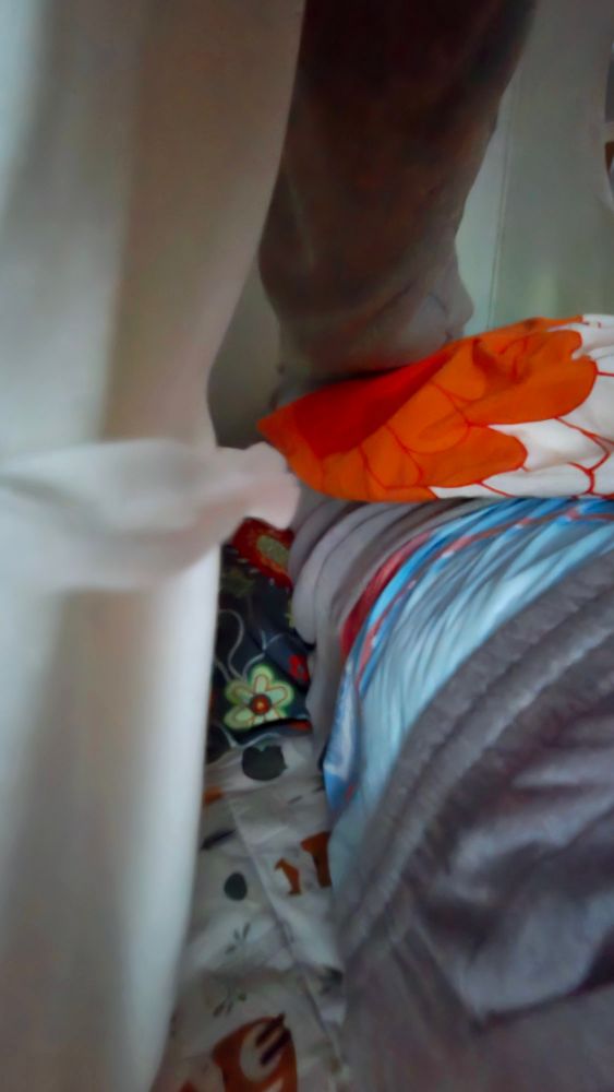A person is lying on their side with a view of their back. They are wearing gray pyjamas, and the bottoms are slid down, revealing a blue diaper with a red waistband. The scene is a cozy bed setup in a tent with multiple layers of blankets and pillows. There is a white curtain on the left side, partially covering the view. The bedding includes a colorful floral-patterned blanket, a blue and gray quilt, and an orange pillow with a white and orange design. The overall atmosphere suggests a comfortable and inviting sleeping area.