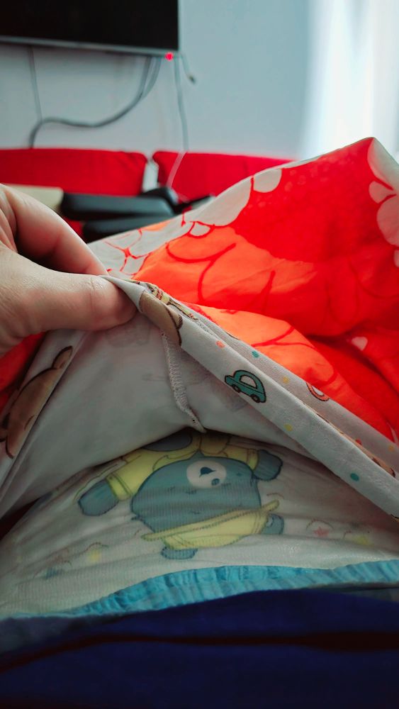 A hand is holding up a section of a blanket or comforter and the front of their pyjama pants. The blanket is bright orange with a floral pattern, while the pyjamas underneath has a light background with small illustrations, including a car and a bear. Under the pyjamas, a diaper is visible with a cartoon bear wearing a yellow outfit. In the background, there is a television mounted on the wall, with visible cables, and a red couch.