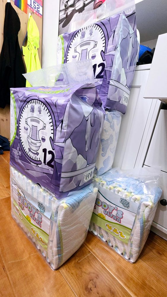 Several packs of diapers are stacked on a wooden floor. The top packs are in purple packaging with a large clock design and the number 12 prominently displayed. Below them are packs in clear packaging showing the diapers inside, with the brand name "KIDDO Diapers" and the product name "Teddy's Ultra" visible. The room has a wooden floor, and in the background, there are clothes hanging, including a bright yellow rain jacket and a black hoodie. A colorful race bib with the number 152 is also visible on the wall.