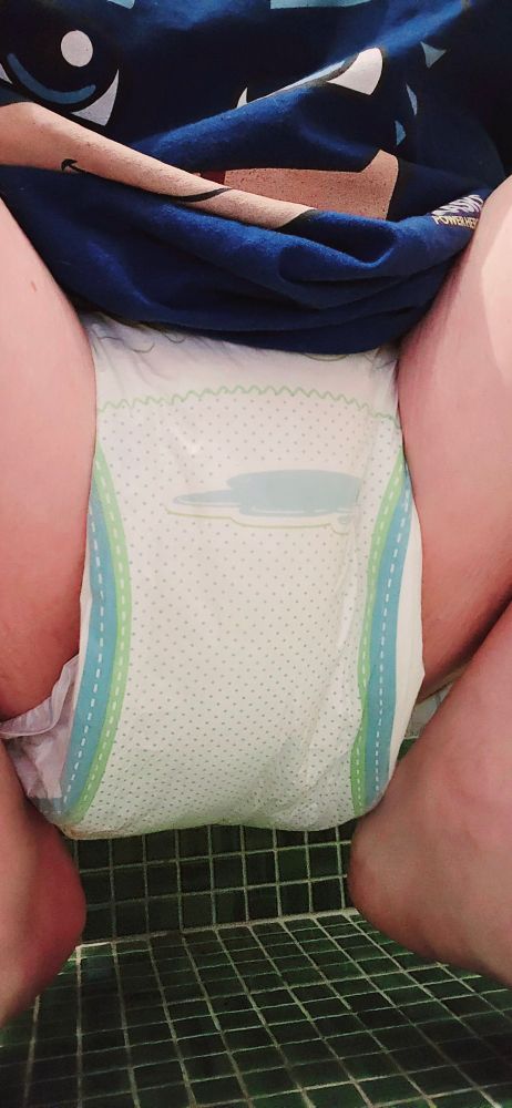 A person wearing a diaper is squatting against a wall with green tiles. The diaper has a pattern with light blue and green lines and dots. The person is wearing a blue shirt with a design that includes white and blue elements.