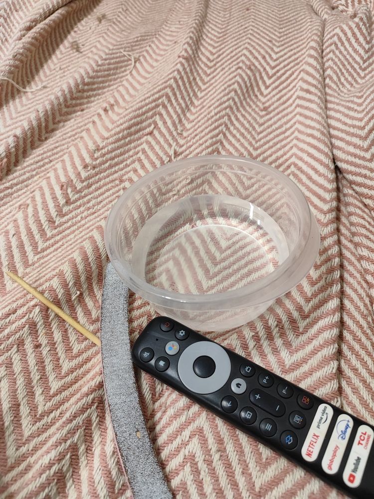 Um potinho transparente com água em cima do sofá, próximo tem um palito, uma lixa de unha e um controle remoto da televisão. 