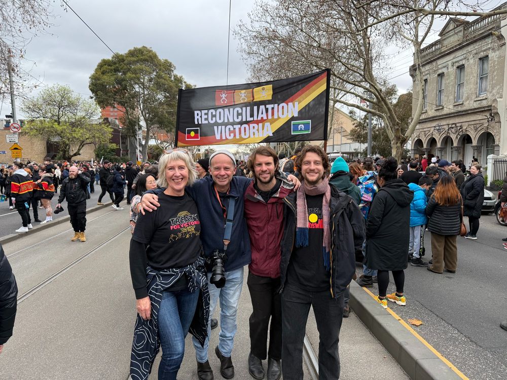 Four people pose and smile together in the foreground of a busy street march. Behind them is a large banner that reads "Reconciliation Victoria" with the Aboriginal and Torres Strait Islander flags. 