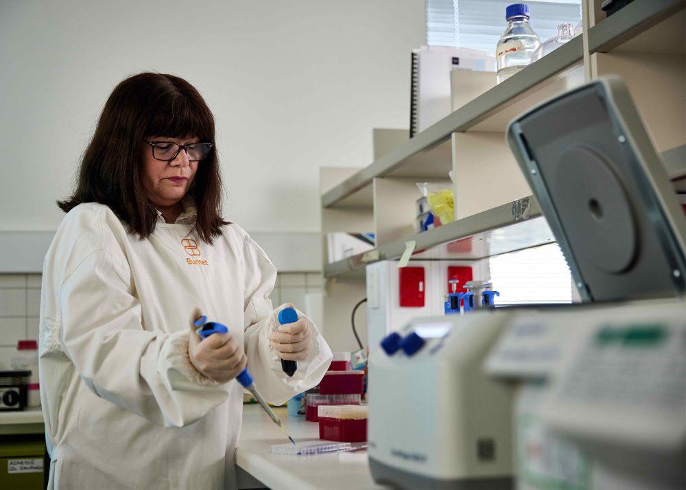 A female scientist wearing a white Burnet-branded lab coat and gloves works in a laboratory. She is using a pipette to dispense liquid into a microplate. 