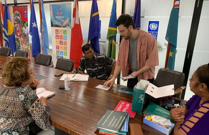 Dean Cassano from Burnet stands and presents printed materials to three seated colleagues during a meeting in Fiji. Behind them are Pacific nation flags and UN Sustainable Development Goals posters.