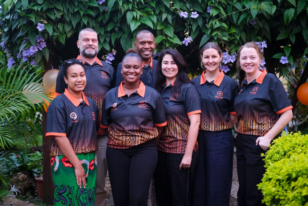 A group of seven Burnet Institute staff members standing outdoors in front of a lush green tree with purple flowers. They are smiling and wearing coordinated black, orange, and patterned shirts with Burnet and Vanuatu logos.