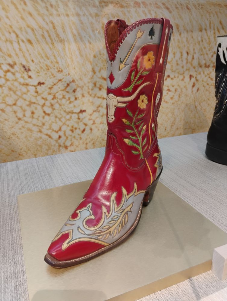 cowboy boot owned by gene autry on display. it is mostly red leather with light blue inlay, as well as stitched pieces of a cow skull, arrows, plants, suits from playing cards. 