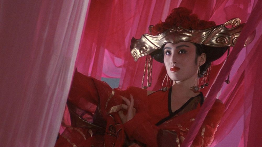 a woman dressed in red and gold robes, with a large gold headpiece dressed with roses. she is surrounded y pink and red chiffon drapes. 
