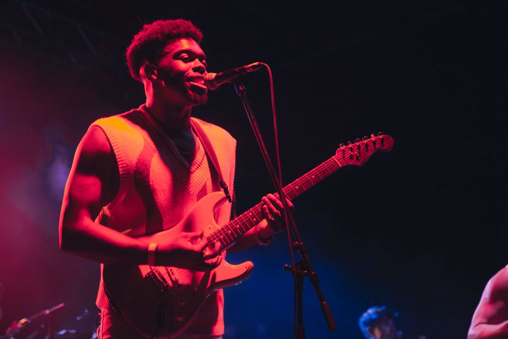 Musician playing electric guitar and singing into a microphone under red stage lighting during a live performance.