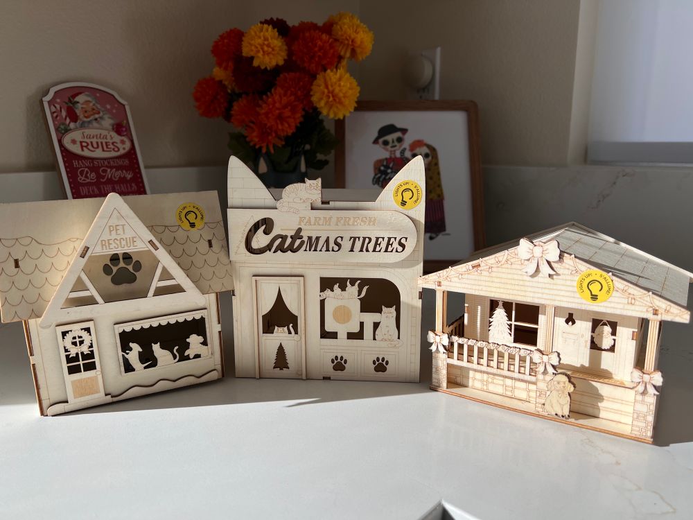 Small Unfinished Wooden LED lit Christmas village buildings. The first one is a pet rescue with 2 dogs and a cat by the window. The 2nd is a farm fresh Catmas trees storefront shaped like a cat’s head with cat trees and cats in the window. The third building is a vintage style home with a retro poodle and bows. 
