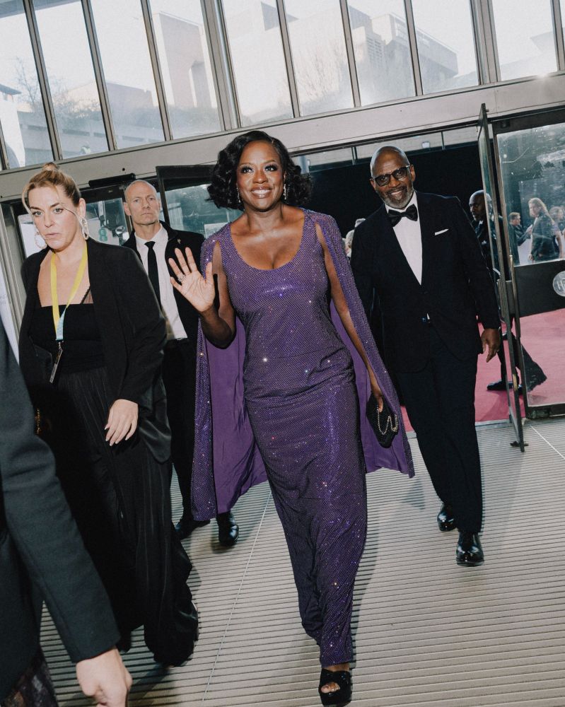 Viola Davis waves as she walks into the EE BAFTA Film Awards. She is wearing a floor length purple sequined dress, her short black hair is curled and she is smiling brightly.