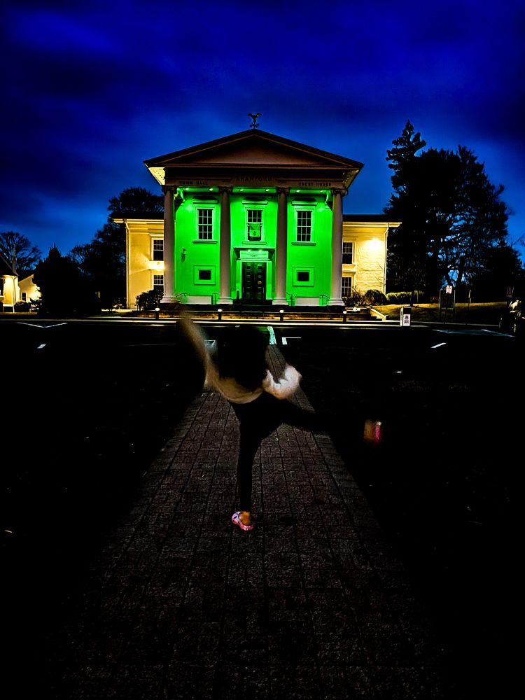 A picture of a town hall light brightly in green with a deep rich blue sky looming above. In the foreground, a child prances. 
