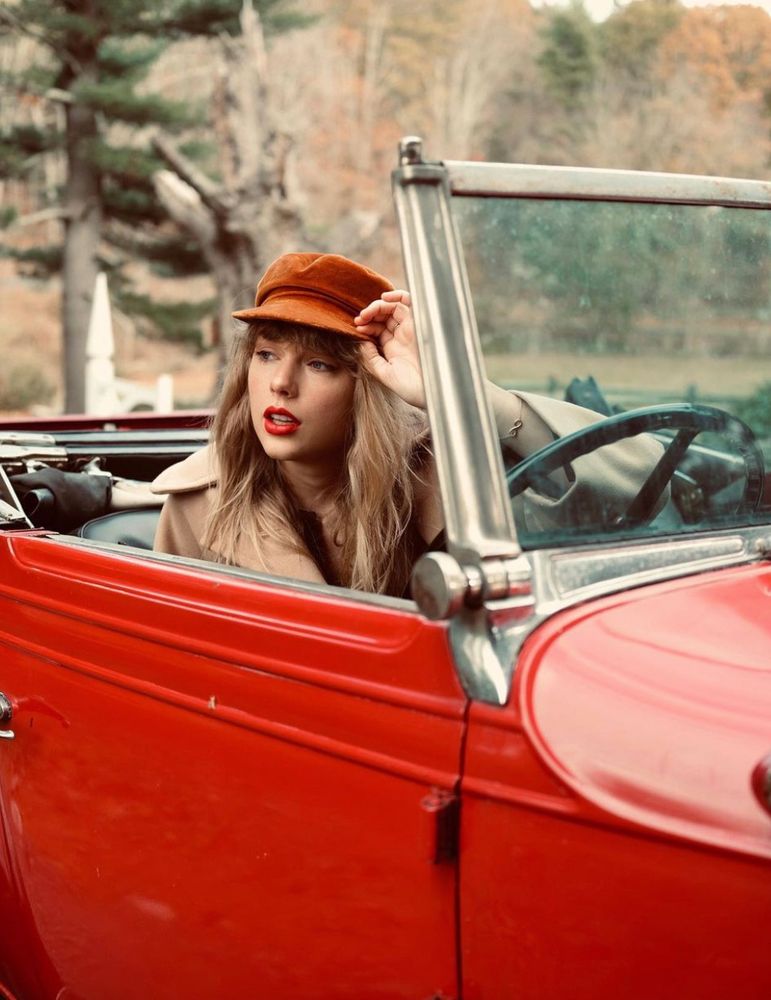 Red TV era Taylor Swift. She is pictured in a red vintage convertible. She is wearing a rust colored cap, with her signature red lip. She has a beige coat on and is holding the bill of her cap. 
