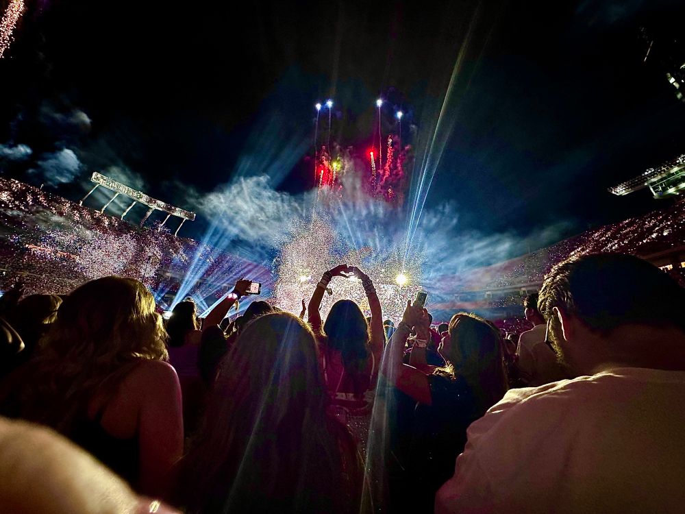 A photo from the crowd at KC N2 of the eras tour. A girl is doing the heart hands symbol as fireworks go off. 