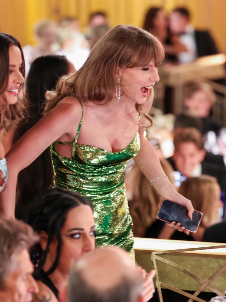 Taylor inside at the GG. She is walking with Keleigh Teller and laughing while holding her phone. 