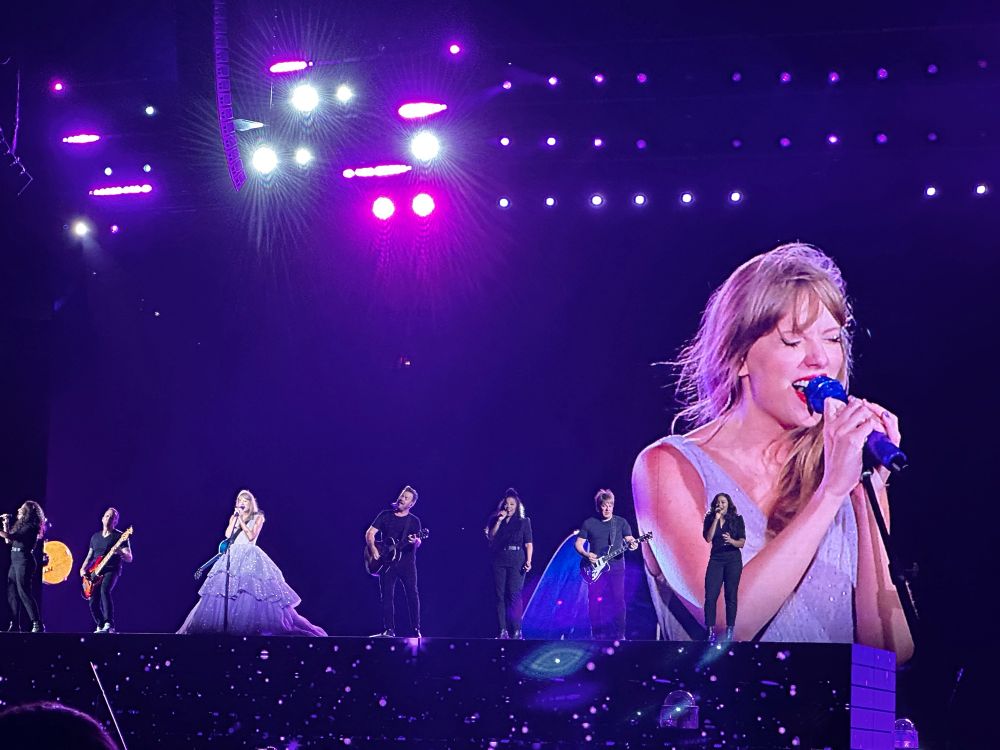 Taylor Swift and her band on stage at the eras tour. She is using the koi fish guitar and surprised the crowd with Long Live for the first time on the tour. She’s wearing the purple cupcake dress. Her band is on stage with her as an homage to the speak now tour, where they did this nightly. 