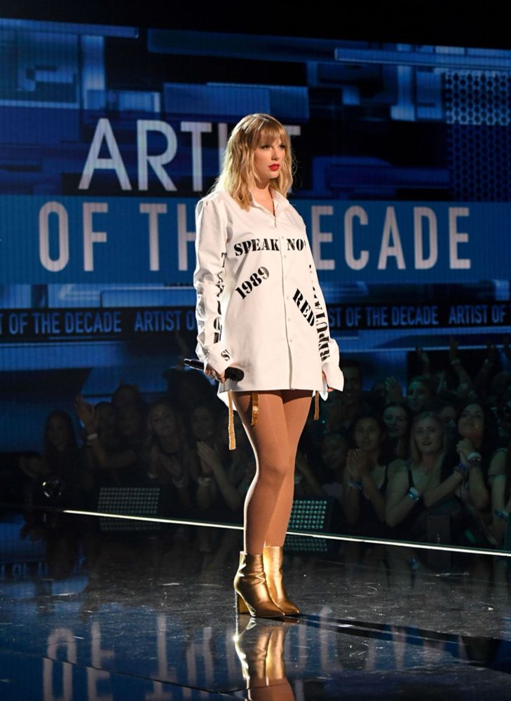 Taylor Swift on stage performing at the AMAs. She is standing in front of the “Artist of the Decade” banner on screen, as she wears the iconic stolen album long sleeve white dress. Golden straps from her bodysuit can be seen, matching her golden boots. 