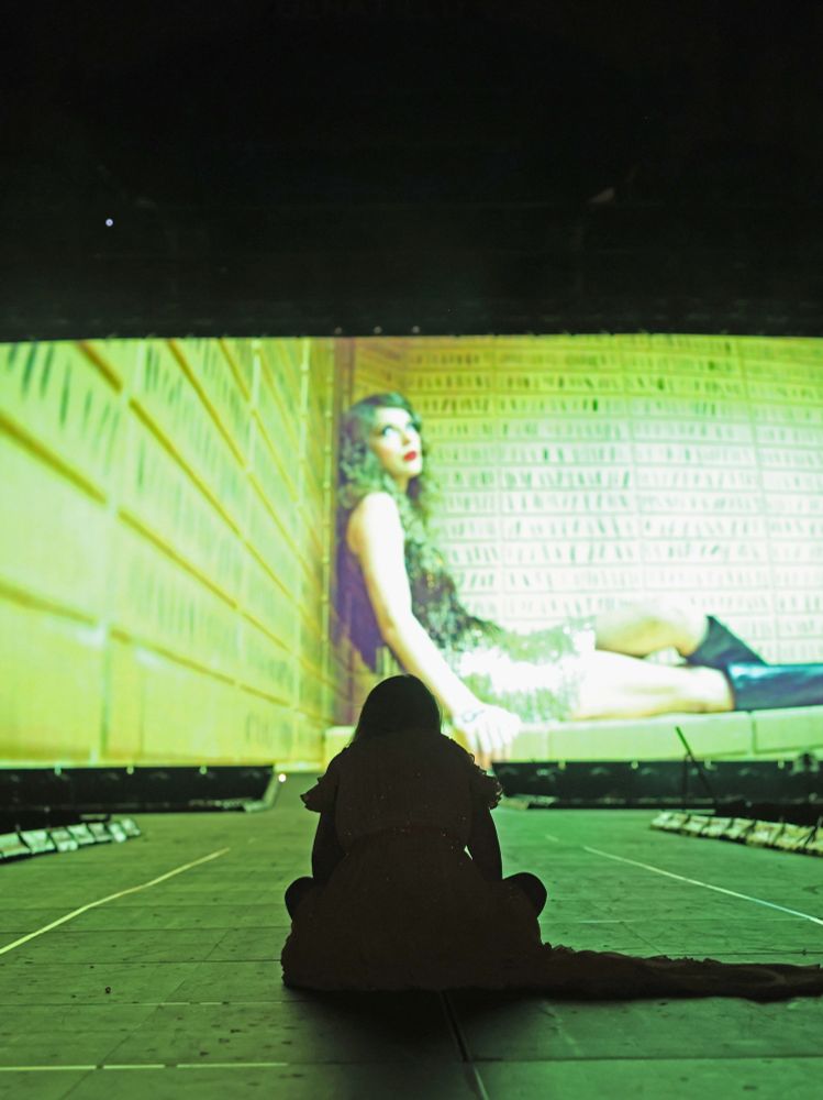 Speak Now TV era Taylor Swift. She is on stage at The Eras Tour, Night 1 in KC, MO. She’s is seated criss-cross on the floor of the stage facing the big screen. She is in the original yellow surprise song dress. She’s watching the premiere of the ICSY music video with the crowd. 