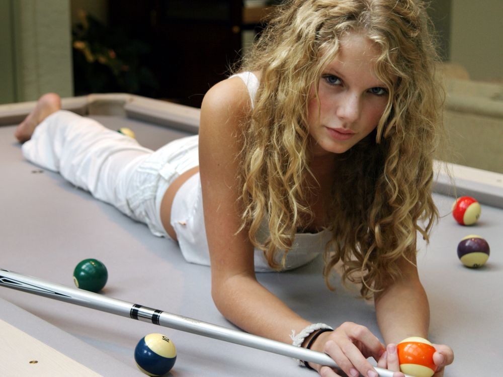 Young Taylor Swift photographed laying on her stomach on top of a pool table. She is wearing white pants and a white tanktop. She’s holding a pool ball with an orange stripe in her left hand and holding the pool stick in her left hand. She has her natural curls down and wearing minimal makeup. 