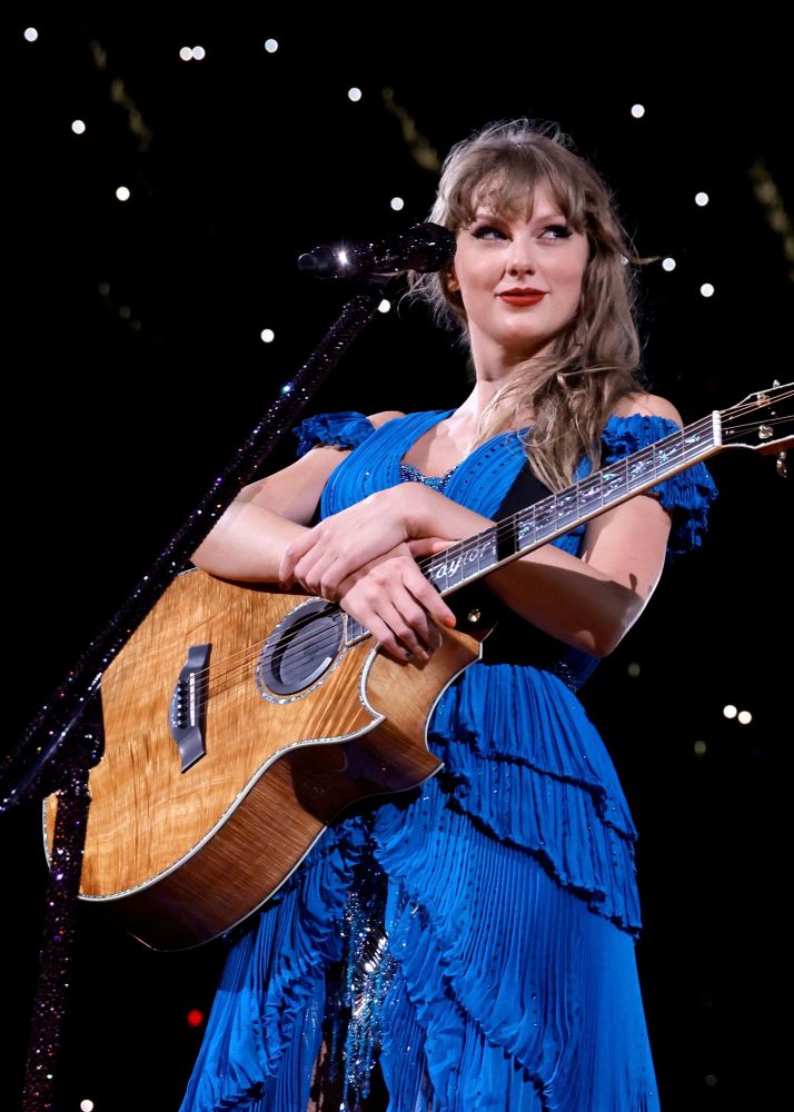 1989 TV era Taylor Swift. She is on stage in LA at The Eras Tour, about to start the acoustic set. She debuted the original blue 1989 set, as well as several other blue outfits throughout the night. She is pictured in the original blue dress worn over the 1989 set. She is holding her guitar and smirking at the crowd. 