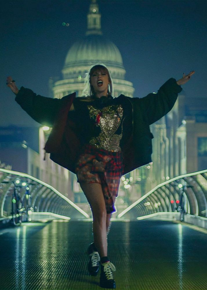 Taylor Swift in the End Game music video. She is walking toward the camera on a bridge. Her arms are extended out and she’s singing. Her outfit is busy, with sequins and plaid and color all over. She has a black jacket and black sneaker heels on. 