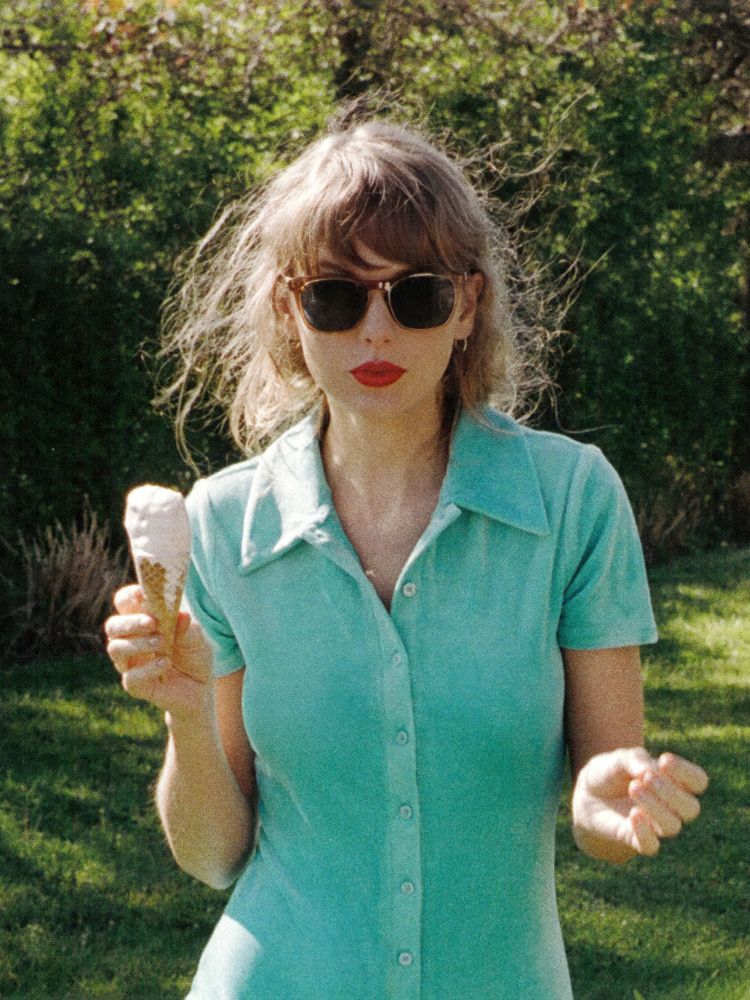1989 TV era Taylor Swift. She is pictured during the 1989 TV album photoshoot, in front of trees while standing on grass. Shes in a blue collared dress. Shes holding a melting ice cream cone. Her hair is in an updo making it look short, and she’s wearing her iconic red lipstick with black sunglasses. 