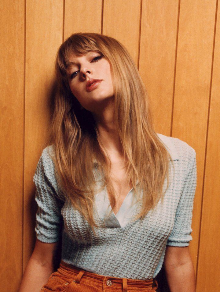 Midnights era Taylor Swift. A photo from the Midnights album photoshoot. Taylor is in front of a wood panel wall. She is wearing a crochet blue top and burnt orange corduroy pants. Her hair is wavy and down as she looks into the camera. 