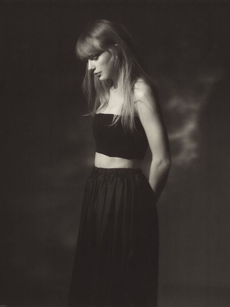 TTPD era Taylor Swift. Pictured for the TTPD album photoshoot, Taylor’s standing in a black and white toned photo. She’s in a two piece black dress outfit and looking down at the ground. 
